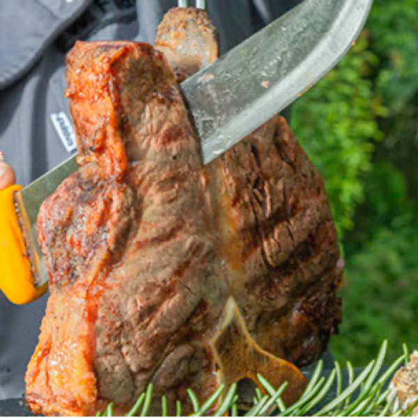 Gro&szlig;es gegrilltes T-Bonesteak, das mit einem Messer geschnitten wird, im Freien mit gr&uuml;nem Hintergrund.