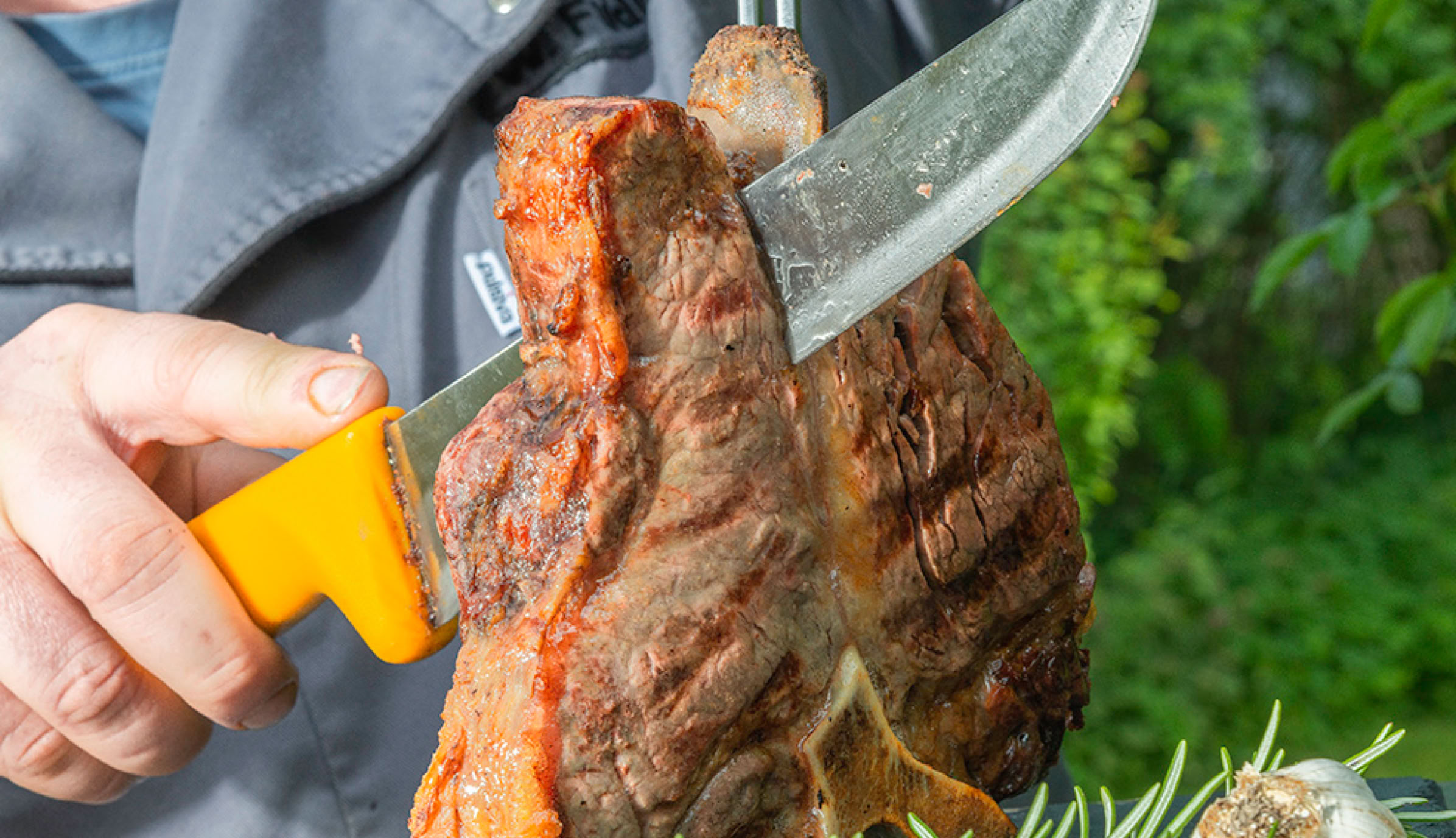Wolfgang Fuldner schneidet ein gro&szlig;es St&uuml;ck gegrilltes Fleisch mit einem Messer, im Vordergrund ist Rosmarin zu sehen.