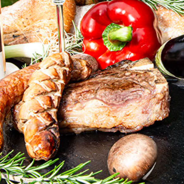 Gegrilltes Fleisch, W&uuml;rstchen, rote Paprika, Rosmarin und ein Pilz auf einem Tisch.